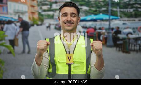 Giovane ispanico in giubbotto riflettente che fa volontariato sulla terrazza del ristorante con posti a sedere all'aperto in una zona vivace, esprimendo entusiasmo e positività, s Foto Stock