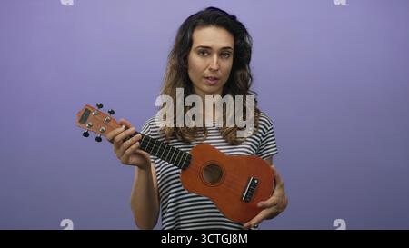 La donna suona ukulele in studio viola con uno sguardo concentrato e una postura rilassata, accordi che premono le dita sulla lavagna; serenità. Foto Stock