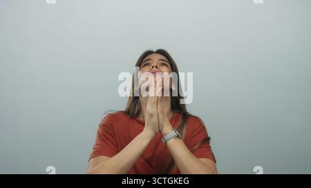 Giovane donna ispanica che prega a occhi chiusi e mani agganciate, in piedi su un isolato sfondo bianco, trasmettendo fede e serenità durante la wearin Foto Stock