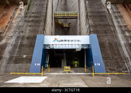 Itaipu Binacional, Brasile - 22 giugno 2025: Il cancello di accesso centrale e la segnaletica Itaipu Binacional sotto il massiccio muro della diga della centrale elettrica. Foto Stock
