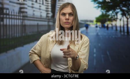 Una giovane donna bionda determinata punta il dito indice verso la macchina fotografica su strade urbane soleggiate; sicurezza. Foto Stock