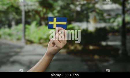 Mano che regge la bandiera svedese in un ambiente cittadino all'aperto che simboleggia l'orgoglio nazionale e la cultura. Foto Stock