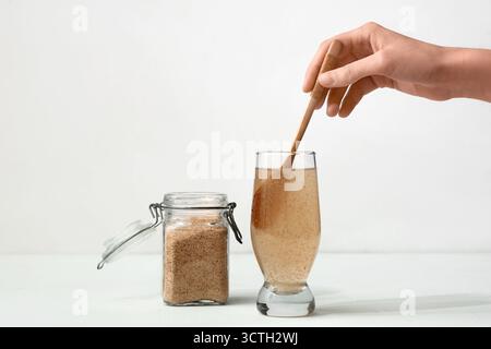 Mano femminile con cucchiaio e buccia di psico in un bicchiere d'acqua su sfondo chiaro Foto Stock