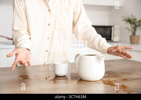 Donna con tazza, caffettiera e caffè versato sul tavolo in cucina Foto Stock