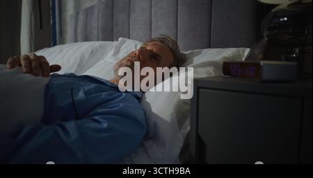 Uomo di mezza età si sveglia presto la mattina in una camera da letto piena di luce solare. L'uomo dai capelli grigi si allunga nel letto, indossa gli occhiali. Concetto di routine mattutina, relax, comfort, mattinata accogliente. Foto Stock