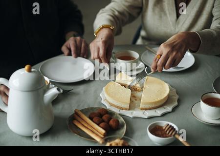 Mani di una donna anziana che affetta cheesecake mentre beve il tè con gli amici seduti accanto a lei a tavola con teiera, tazze, grissini, tartufi, miele e piatti vuoti durante la riunione Foto Stock