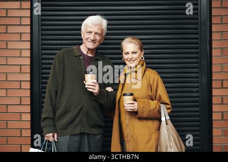 Uomo e donna caucasica senior in piedi all'aperto tenendo tazze da caffè da asporto, sorridendo davanti alla macchina fotografica, uomo che porta borse della spesa, donna che collega le braccia, coppia a piedi Foto Stock