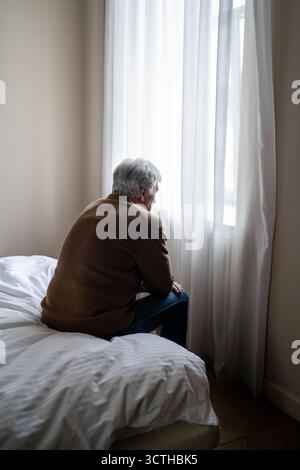 Uomo anziano seduto da solo in condizioni di affaticamento mentale e profonda depressione da finestra, retrovisione Foto Stock