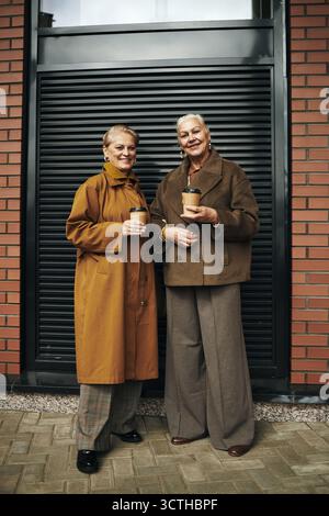 Due donne caucasiche anziane in piedi all'aperto che tengono tazze da caffè da asporto, sorridono e guardano la macchina fotografica, indossano giacche autunnali, in piedi di fronte al moderno edificio in mattoni Foto Stock