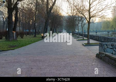 Vicolo del parco cittadino con panche e alberi lungo il fiume alla luce del mattino presto. Tranquilla scena urbana che simboleggia il relax, la tranquillità e la connettività Foto Stock