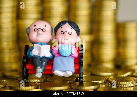 Statuette di coppia anziane in miniatura sedute sulla panchina con pile di monete d'oro, che rappresentano la pensione, i risparmi, i fondi pensione e la sicurezza finanziaria Foto Stock