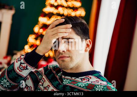 Giovane uomo in maglione natalizio al chiuso, che sente lo stress delle vacanze vicino all'albero di Natale splendidamente illuminato Foto Stock