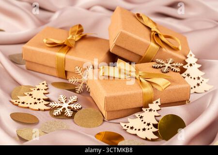 Scatole regalo marrone con nastri dorati su sfondo di seta. Scatole regalo, abeti di Natale in legno, fiocchi di neve e coriandoli rotondi. Foto Stock