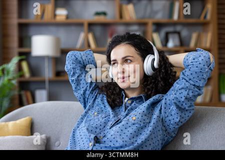 Una giovane donna sorridente con capelli ricci che si rilassa su un comodo divano di casa, si diverte mentre ascolti l'audio tramite le cuffie wireless Foto Stock