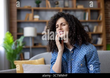 Giovane donna che sente un intenso dolore dentale, toccando la guancia e la mascella, soffre di un forte mal di denti mentre siede su un divano in un interno di casa, che necessita di cure mediche immediate Foto Stock