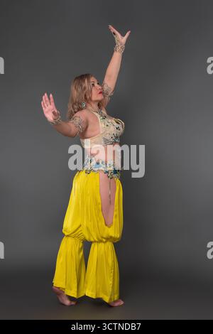 Vista laterale di una donna adulta di mezza età con braccia allungate e alzate che indossano un abito da ballo: La parte superiore e i pantaloni harem gialli si alzano a piedi nudi in lengt Foto Stock