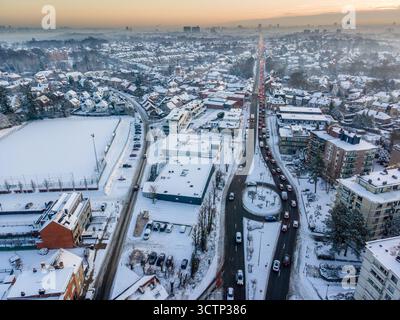 Ripresa aerea di una lunga strada rettilinea a Dilbeek in una bianca mattinata d'inverno, neve che copre la città su entrambi i lati, che conduce verso il lontano skyline Foto Stock