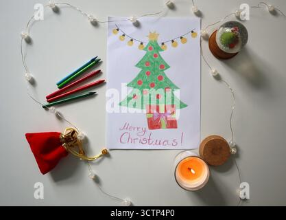 Piatto di alberi di Natale disegnati a mano con regalo e testo di buon Natale, circondato da pastelli, candele, globo di neve e luci delle fate. I bambini Chris Foto Stock
