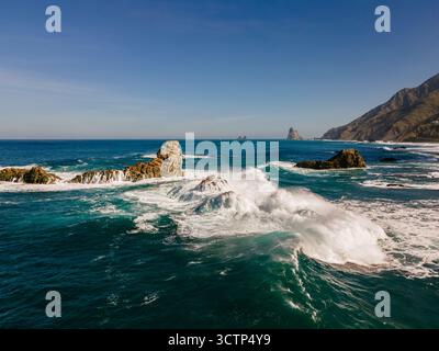 Vista aerea ravvicinata delle onde che si infrangono su aspre rocce vulcaniche sulla costa settentrionale di Tenerife, con spruzzi e schiuma in contrasto con il mare azzurro Foto Stock