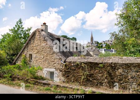 Un affascinante cottage con tetto di paglia si trova accanto a un muro intemprato, con il pittoresco villaggio di Pont-Croix, in Bretagna, e un'alta guglia della chiesa nel backgroun Foto Stock