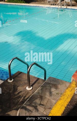 Una vista soleggiata e ad alto angolo di una piscina vuota con maniglie cromate per scala e gradini in pietra testurizzata caratterizzati da una striscia di avvertimento giallo brillante Foto Stock