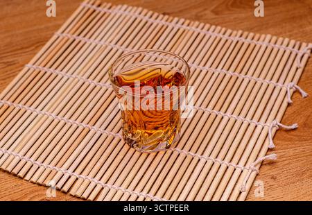 Un bicchiere di bevanda ambrata poggia su un tappeto di bambù posto su una superficie di legno. Creare una natura morta calda e invitante. Foto Stock