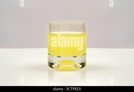 Un bicchiere trasparente riempito a metà con succo giallo rinfrescante, posizionato su una superficie bianca pulita con sfondo neutro. La vivace bevanda spicca. Foto Stock