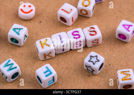 Le perle di lettere bianche che scrivono "KISS" sono disposte centralmente su una superficie testurizzata. Circondato da altre perle dell'alfabeto sparse. Creare un gioco Foto Stock