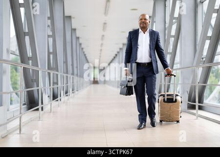 Un uomo nero maturo in tuta che cammina con sicurezza attraverso il corridoio dell'aeroporto e porta valigie Foto Stock