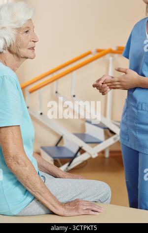 Anziana donna caucasica seduta sul divano nel centro di riabilitazione e guardando in lontananza con attenzione mentre si prepara per la terapia fisica con l'infermiera Foto Stock