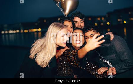 Un gruppo di amici allegri che scattano un selfie all'aperto di notte, illuminato dalle luci della città. Foto Stock