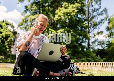 Moderna imprenditrice multitasking telefono cellulare e laptop mentre è seduta sull'erba nel parco cittadino Foto Stock