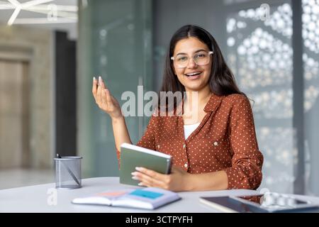 Giovane donna d'affari indiana con occhiali e camicia a pois sorridente e parla a una riunione, tenendo un notebook a una scrivania bianca con libri aperti, atteggiamento professionale amichevole Foto Stock