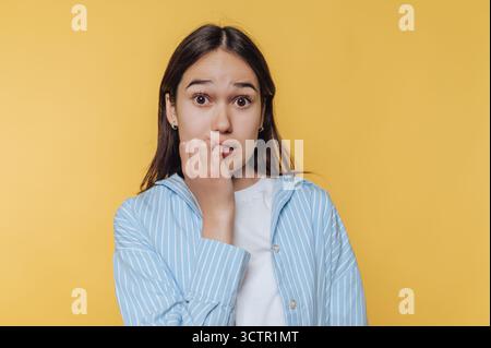 Giovane donna che morde le unghie, con un aspetto ansioso su sfondo giallo. Foto Stock
