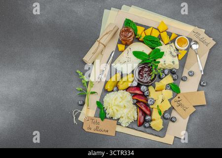 Vassoio di formaggi con Tete de Moine, formaggi assortiti, frutta, marmellata ed erbe aromatiche su un tavolo di ardesia. Sfondo in pietra nera in calcestruzzo, piano, vista dall'alto Foto Stock