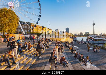 Vista del tramonto della città vecchia di Dusseldorf con la Torre del Reno sullo sfondo. I turisti si godono la calda luce del sole camminando lungo l'argine Foto Stock