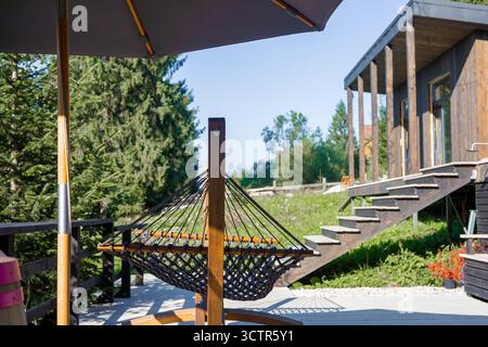 Un moderno spazio esterno con un'amaca, un ombrellone da patio e scale in legno che conducono ad una casa contemporanea circondata dal verde. Foto Stock