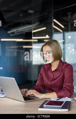 Donna d'affari felice che indossa occhiali e una camicia marrone, concentrandosi sul suo lavoro, scrivendo su un computer portatile a una scrivania di legno chiaro in un ambiente aziendale elegante e sfocato Foto Stock
