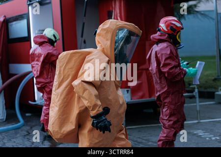 Soccorritori di emergenza in tute antiagenti chimici arancioni e rosse e maschere facciali complete in piedi accanto a un veicolo rosso di risposta a una decontaminazione o. Foto Stock