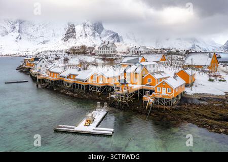La vista aerea delle vivaci cabine arancioni annidate su palafitte incontra le acque frigide, sullo sfondo di montagne innevate, Sakrisøy, Nordland, Norvegia. Foto Stock