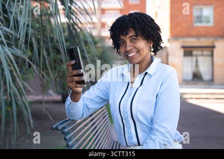 Donna afroamericana con un cellulare che guarda alla macchina fotografica, seduta su una panchina in un parco. Foto Stock