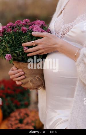 Primo piano bella donna incinta in abito bianco con pizzo che regge un vaso di fiori di crisantemo rosa accanto al bump del bambino. Concetto di aspettativa di maternità Foto Stock
