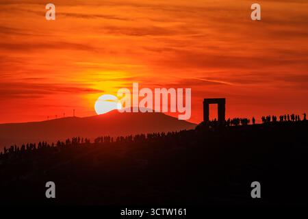 la folla circonda la portara a naxos, l'antico telaio greco della porta si staglia contro il cielo nuvoloso arancione mentre il sole tramonta dietro la collina sullo sfondo Foto Stock