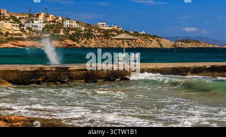 due sedie vuote sotto un ombrellone si trovano su un molo di cemento basso mentre le onde si tuffano in una giornata tranquilla e soleggiata sul mare di un'isola greca Foto Stock