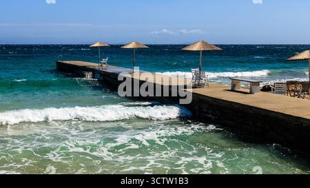 una tranquilla giornata di sole al mare su un'isola greca, una bassa banchina di cemento con tre ombrelloni e sedie vuote raggiunge il mare turchese Foto Stock
