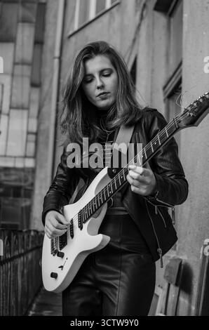 Chitarrista ragazza con giacca in pelle e pantaloni in pelle con chitarra elettrica bianca in stile City Street Rock Foto Stock