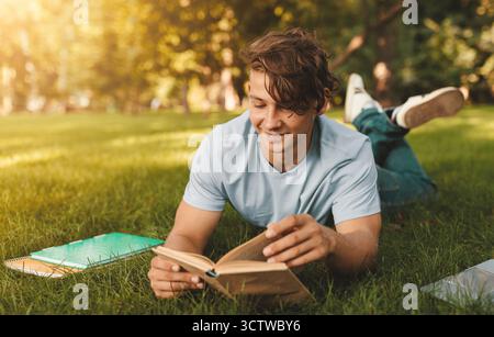 Allegro studente caucasico libro lettura all'aperto, preparando per le lezioni mentre si trova sul prato nel campus universitario Foto Stock