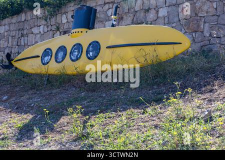 MASLINICA, CROAZIA, 08.25.2025: Una scultura subacquea di colore giallo vivo poggia su un prato e roccioso in primo piano contro un grezzo muro di pietra. Quattro oblò circolari Foto Stock