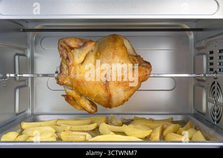 Pollo arrosto succoso e dorato su uno spiedo sopra un vassoio di patate condite in un caldo forno casalingo, invitando a una piacevole cena in famiglia. Foto Stock