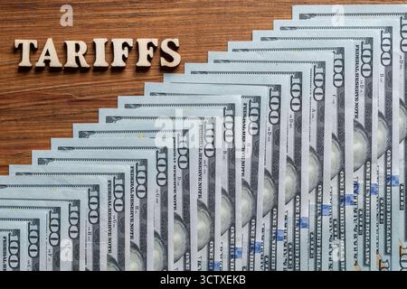Lettere in legno con le tariffe di iscrizione e banconote americane di $100 sul tavolo, vista dall'alto. Tariffe sovrapposte ai dollari americani, simboleggiano le guerre commerciali A. Foto Stock